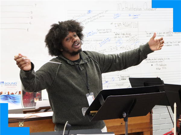  A man holds a conductor baton and gestures widely while leading an orchestra of students.